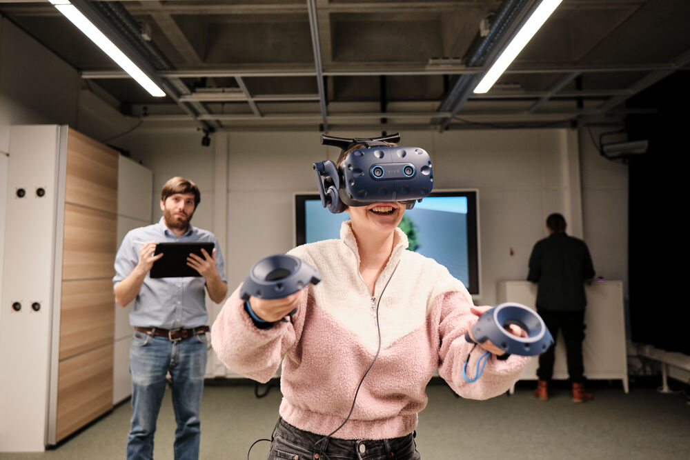 A woman is wearing VR glasses. Behind her is a large screen and two other people.
